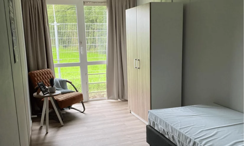 Student room with a chair, bed, closet and table next to a big window